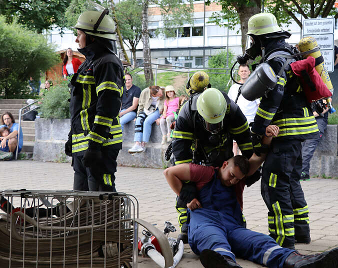 Drei Männer in Feuerwehruniform, einer mit Atemschutzmaske, versorgen einen auf dem Boden liegenden Mann.